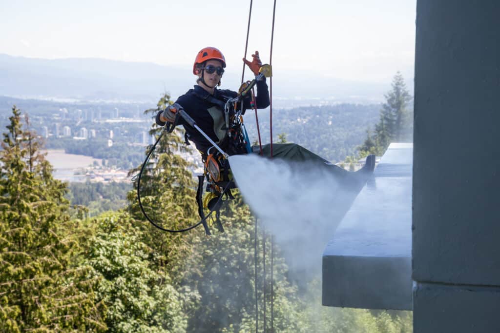 High rise rope access window cleaner is power washing the building during a hot sunny summer day. Taken in Burnaby Mountain, Vancouver, BC, Canada. right professional window cleaner