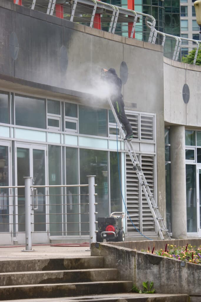 Commercial Pressure Washing. Technician pressure washes the exterior of a commercial building from a ladder.